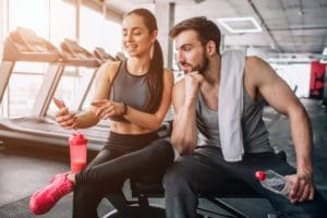 Attractive man and woman sitting on a weight bench and relaxing after long-term exercising while the woman shows something on the phone to her partner.