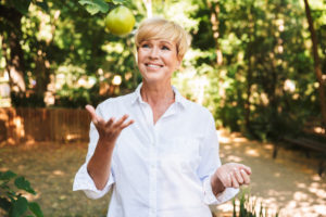 Mature woman tossing up an apple in a backyard.