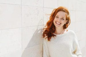Smiling woman with red hair wearing a white sweater leaning against a white wall.